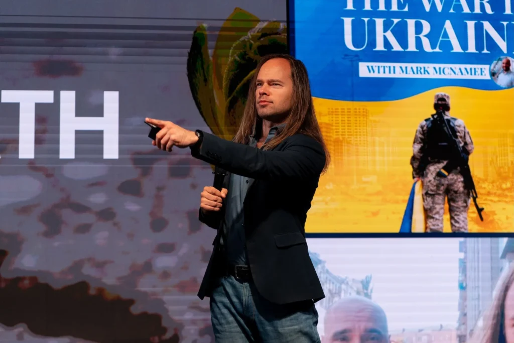 Мерайн Маркус на конференції виступає зі сцени з мікрофоном на фоні українського прапору